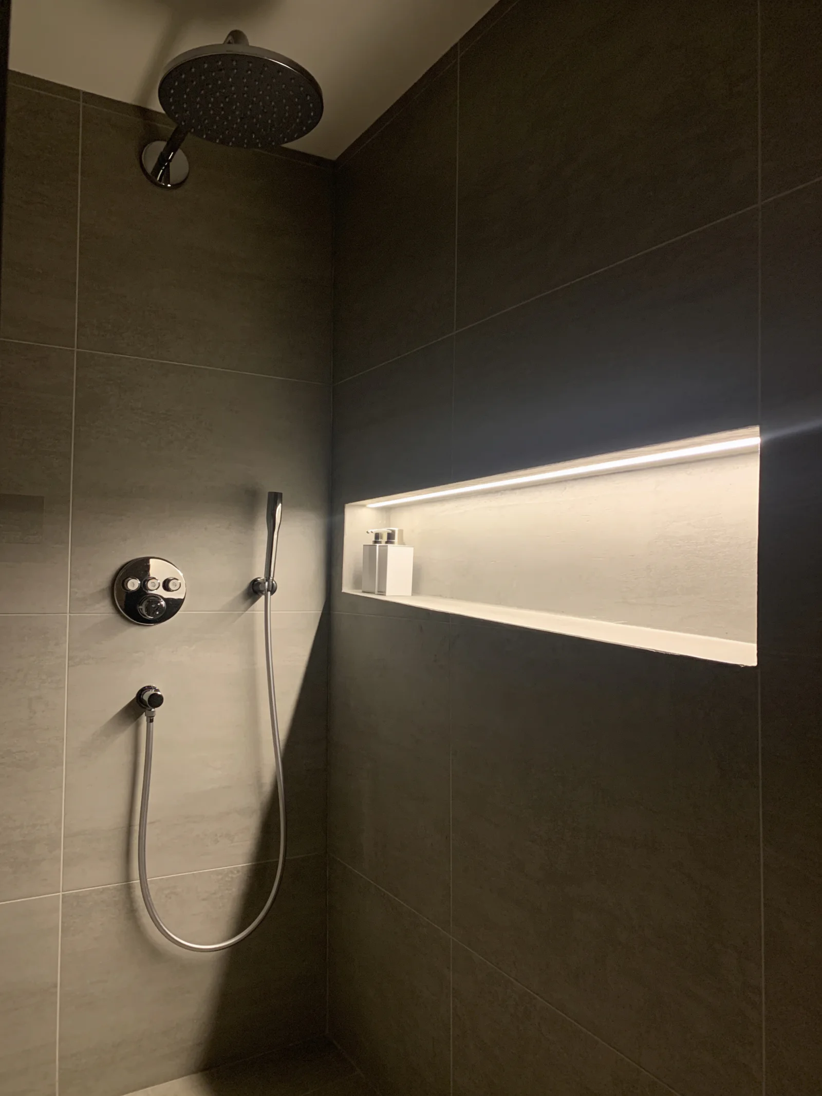 Fertiggestellte Dusche mit dunklen Großformatfliesen, Kopfbrause und beleuchteter Wandnische